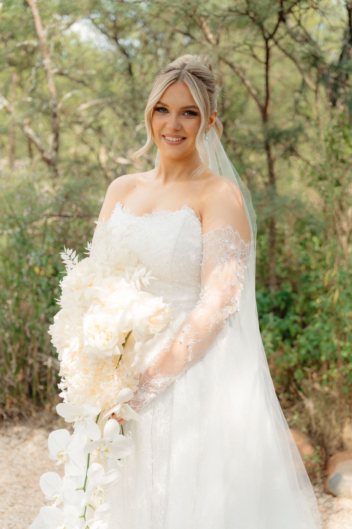 A bride in a white lace gown poses outdoors among trees while holding a white floral bouquet.