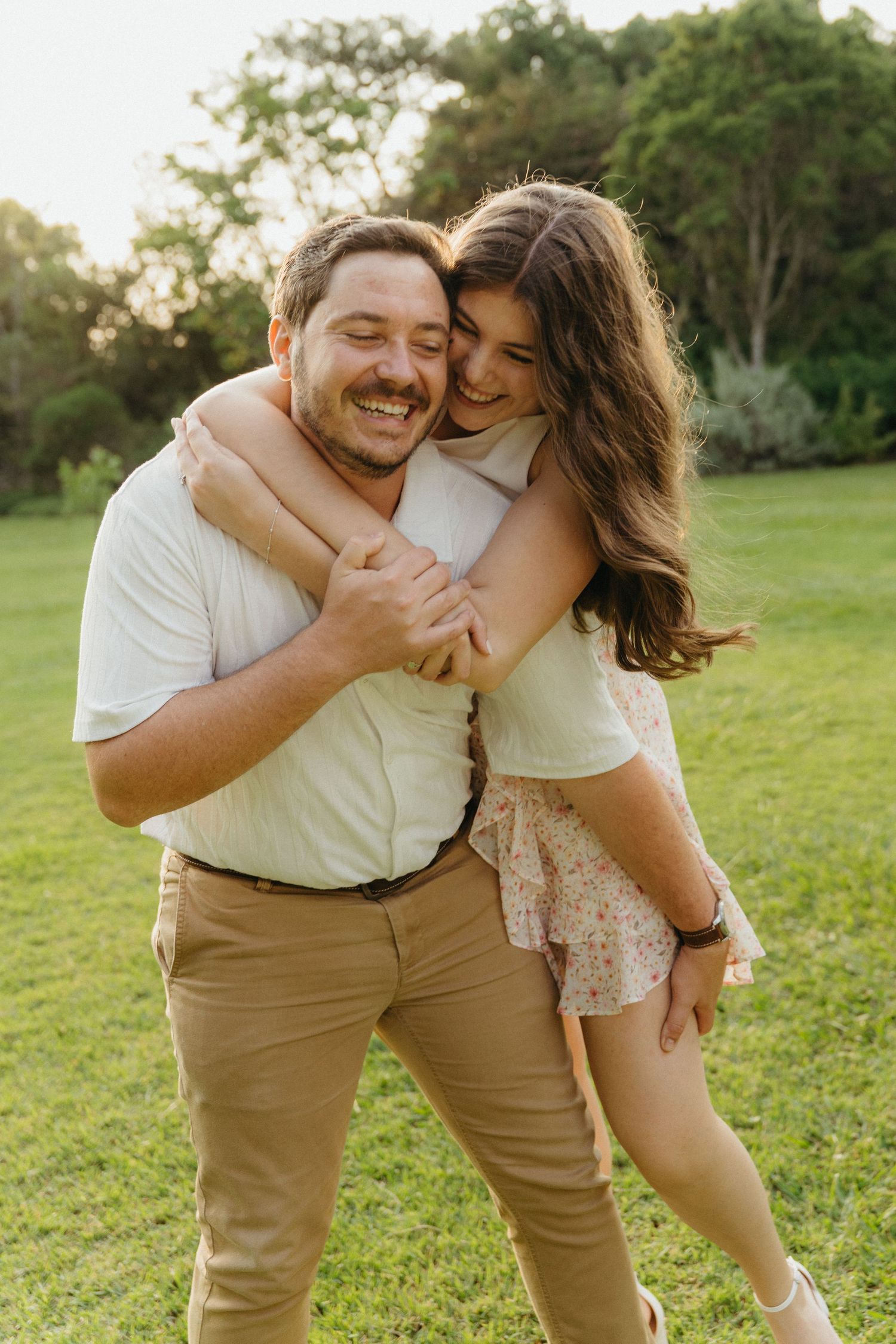 Happy couple sharing a joyful piggyback hug outdoors on green grass with trees in the background.