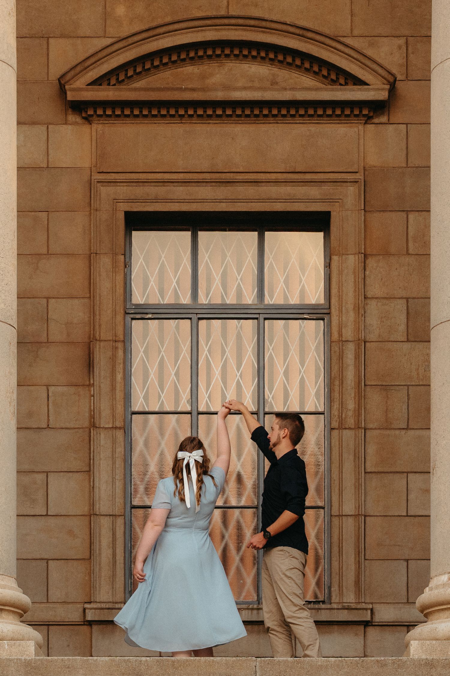 A couple in formal attire poses in front of a stone building with arched windows and columns.