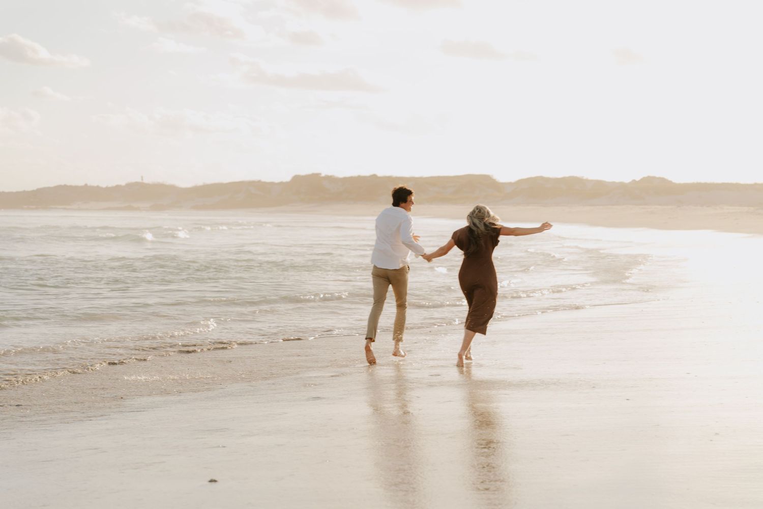 Couple running playfully along ocean shoreline at sunset during romantic Western Cape beach engagement session.