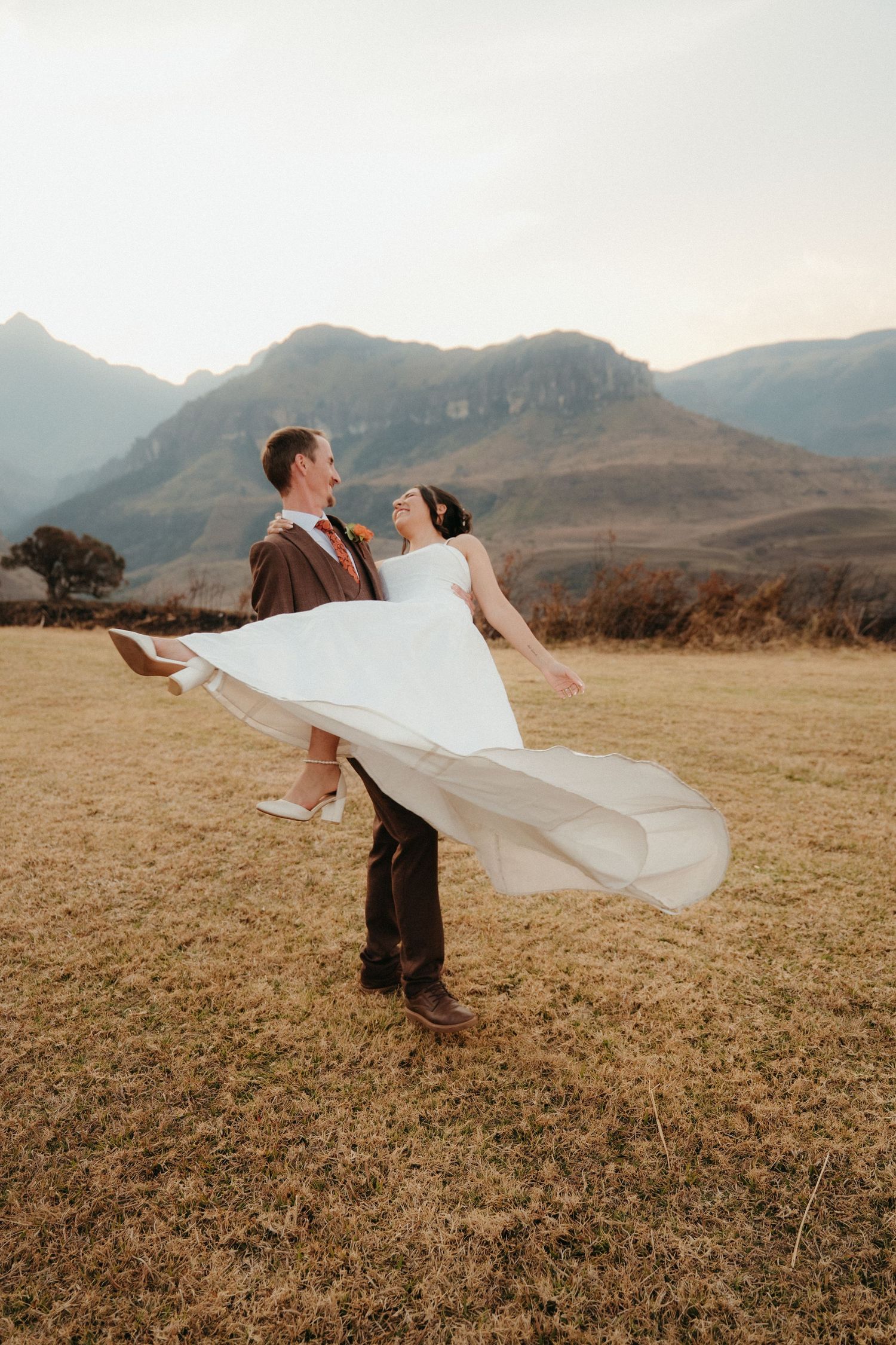 Groom lifts bride in white dress, laughing together in a mountain field in the Drakensberg at sunset during their outdoor wedding.