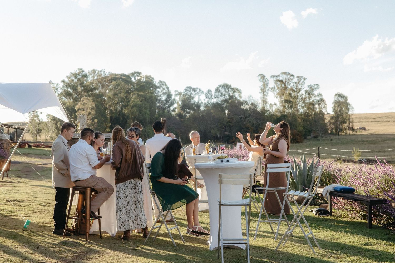 Guests mingle at outdoor cocktail hour tables on a sunny day at a rustic countryside wedding venue.