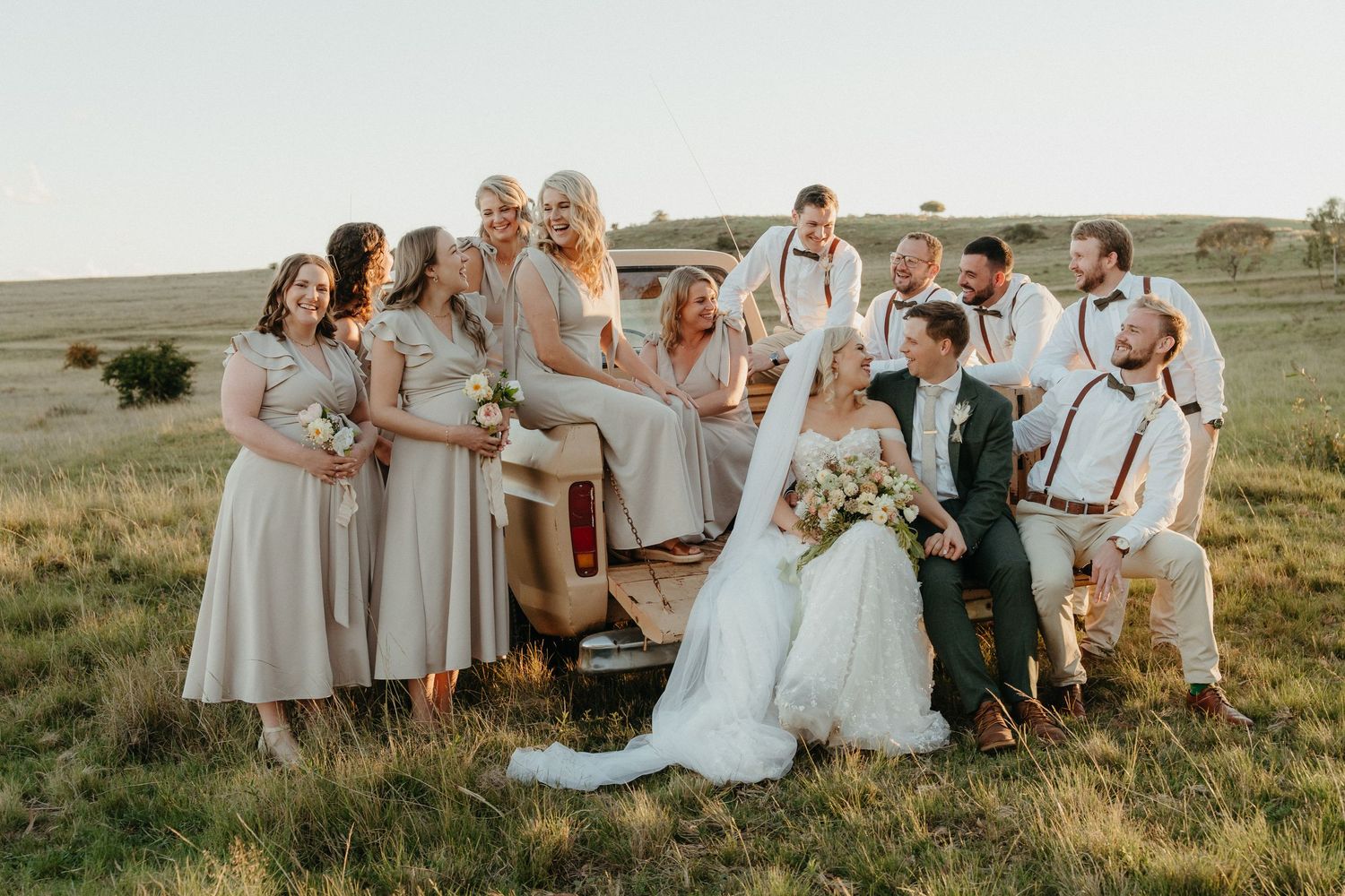 Wedding party poses with bride and groom beside vintage truck in golden field, bridesmaids in champagne dresses, groomsmen in white shirts.