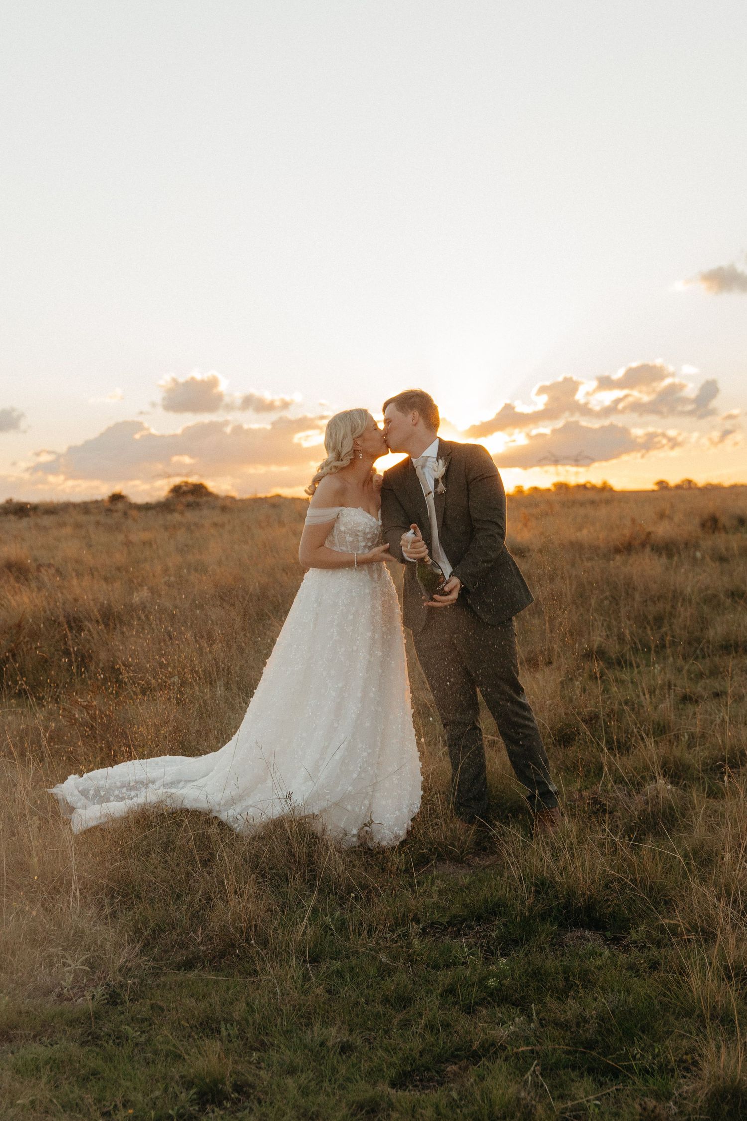 Bride and groom kissing in golden field at sunset, bride in lace gown with train, groom in gray suit.