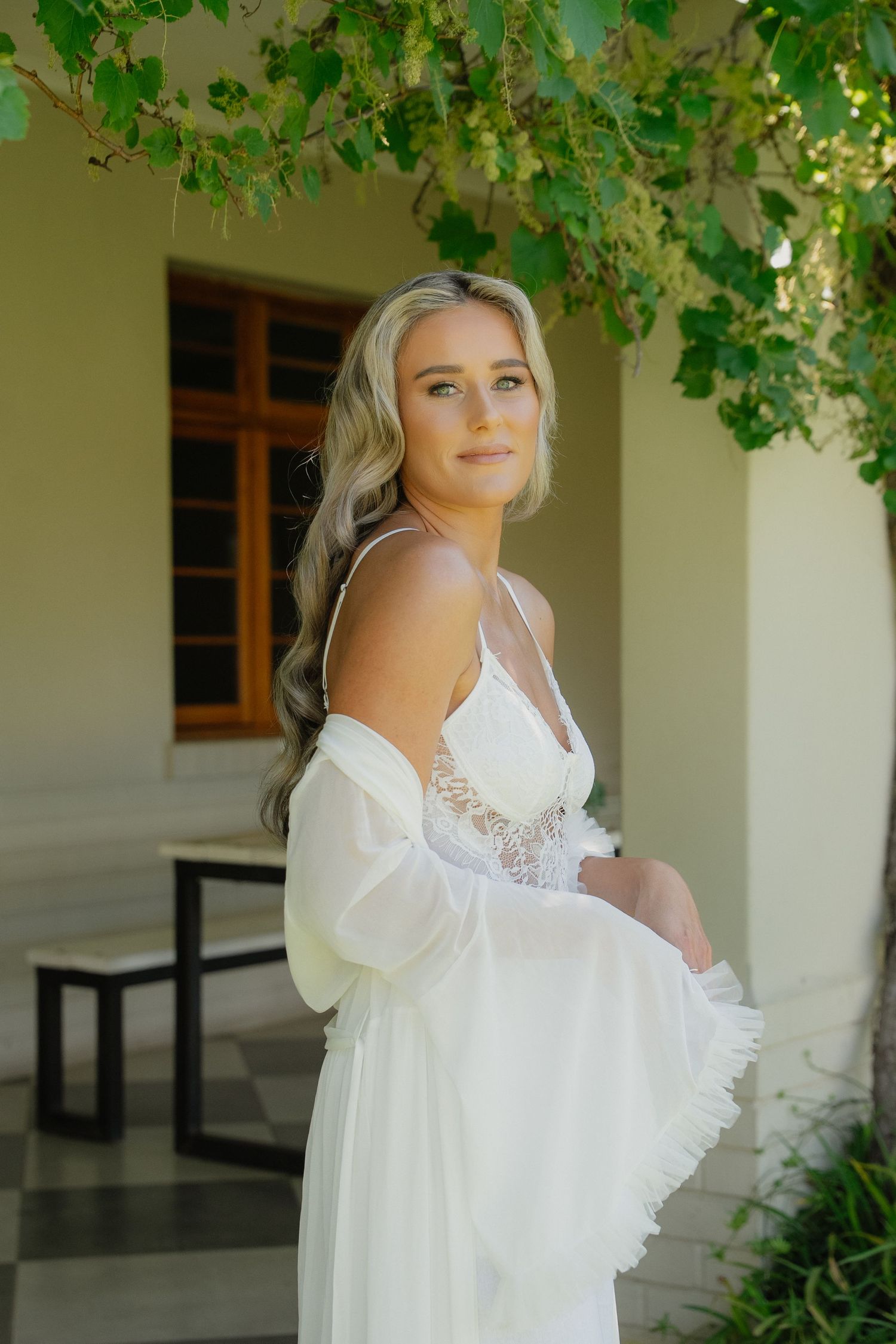 Blonde bride in white lace gown and sheer wrap poses gracefully under green vines at elegant venue.