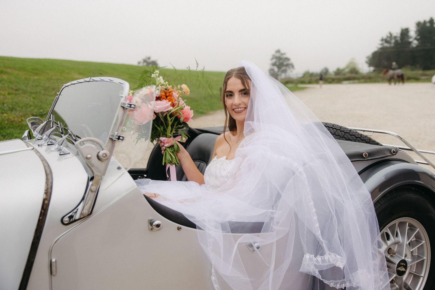 Elegant bride in white veil holding pink bouquet seated in vintage white convertible car on wedding day.