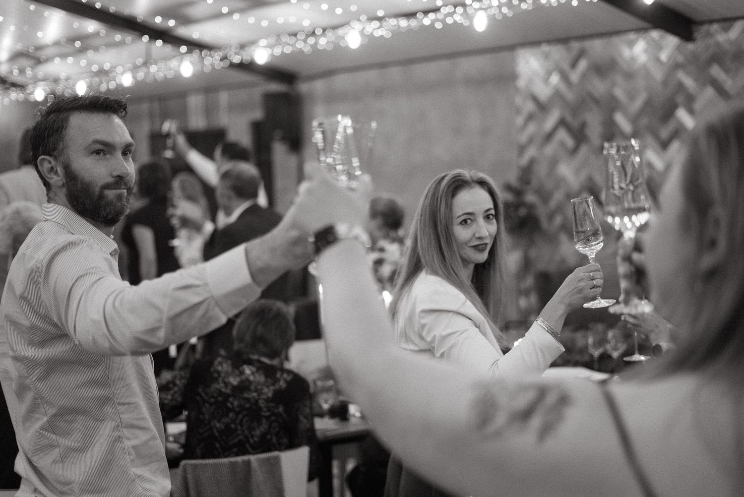 Black and white photo of guests raising champagne glasses in a toast at an elegant indoor celebration.
