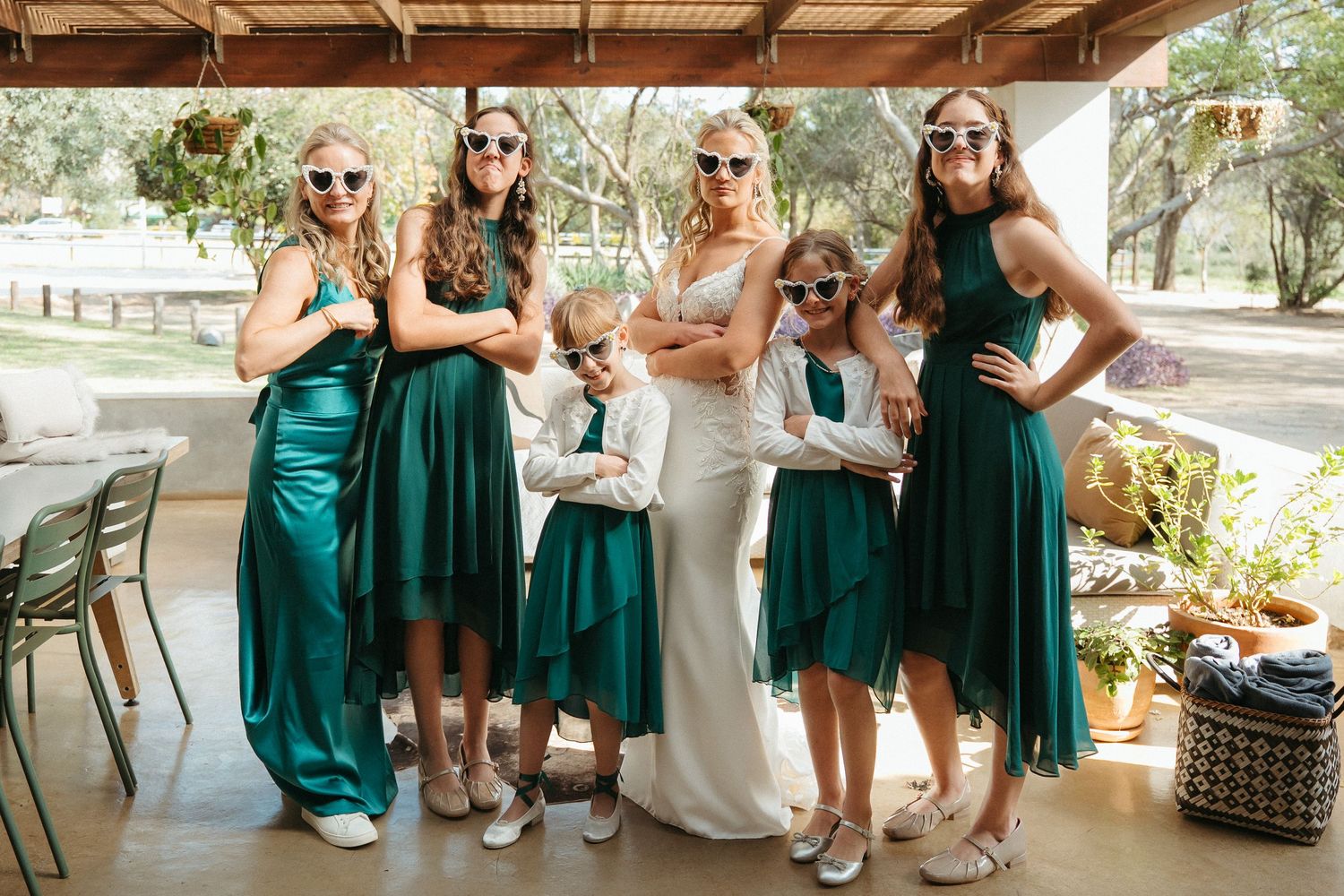 Bride in white gown poses with bridesmaids in emerald green dresses and flower girls wearing fun sunglasses at outdoor wedding.