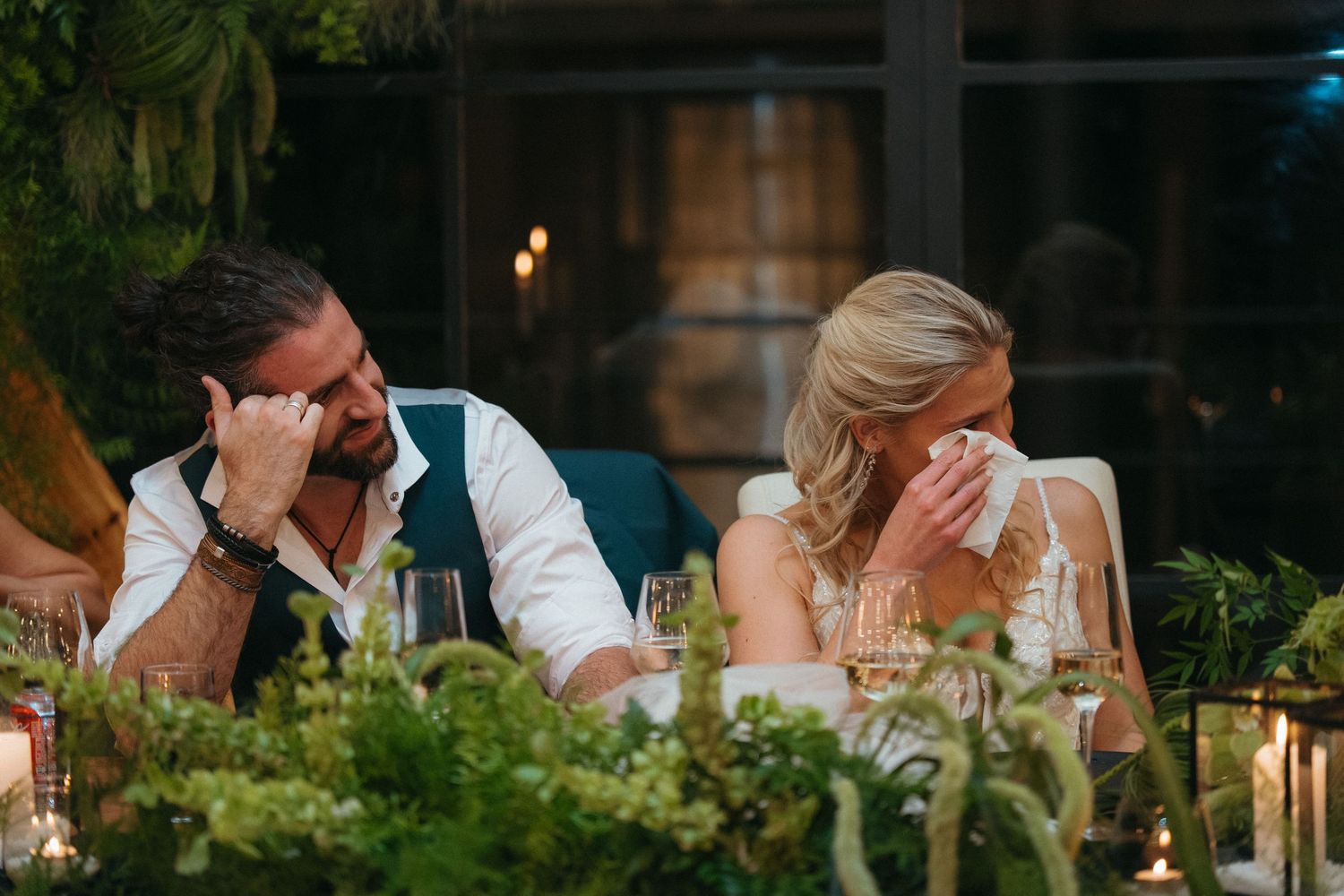 Emotional bride and groom wiping tears during heartfelt wedding reception speeches at a candlelit table with lush greenery.