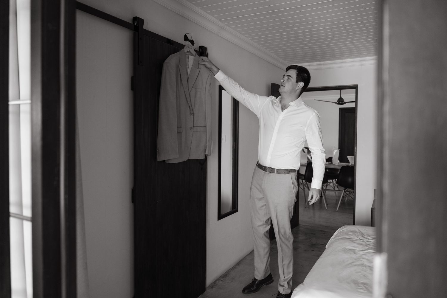 Black and white photo of a man in a white dress shirt and slacks reaching for a suit jacket hanging on a sliding barn door in a bedroom.