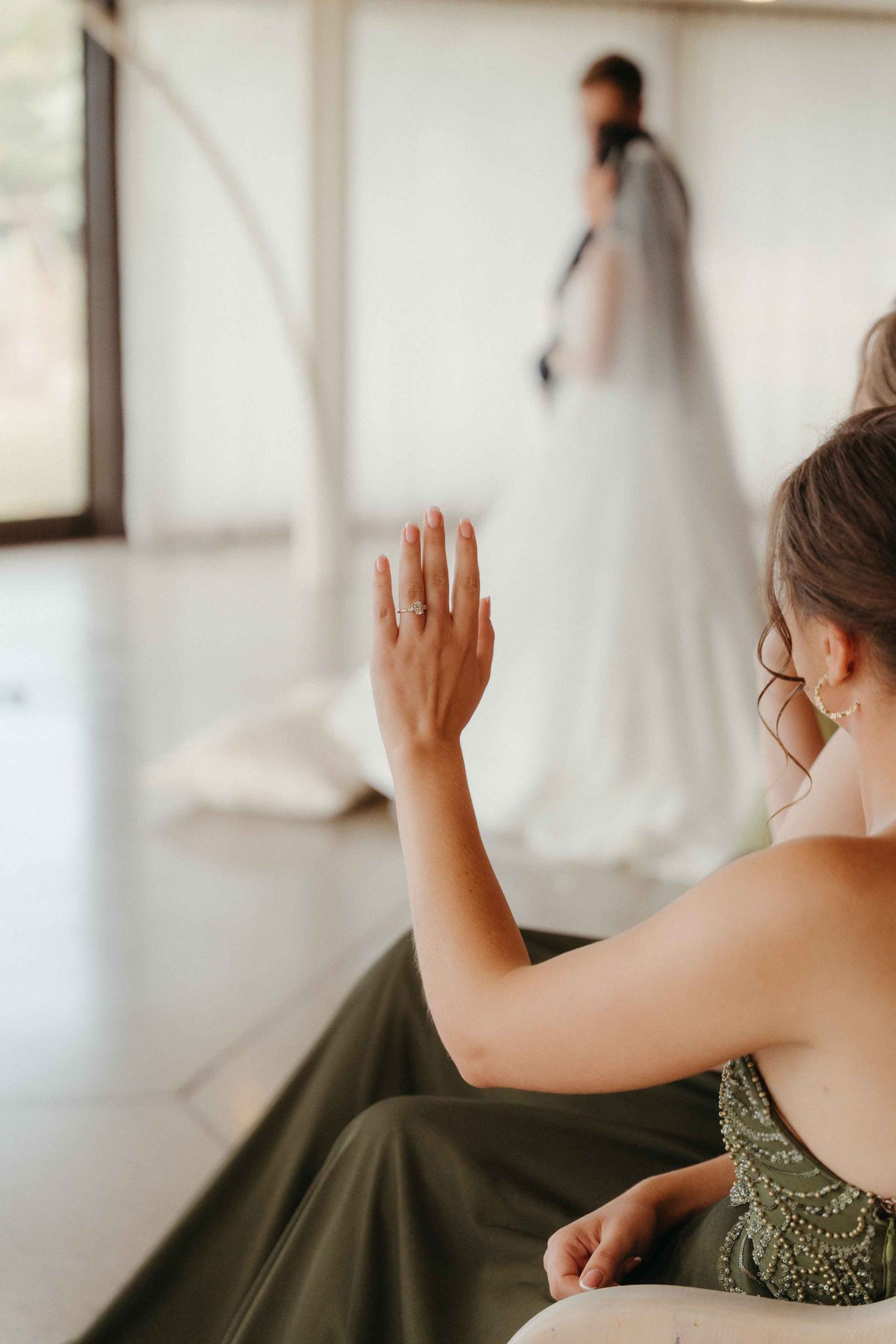 Bridesmaid in green dress raises hand showing ring while bride in white gown and veil stands in background.