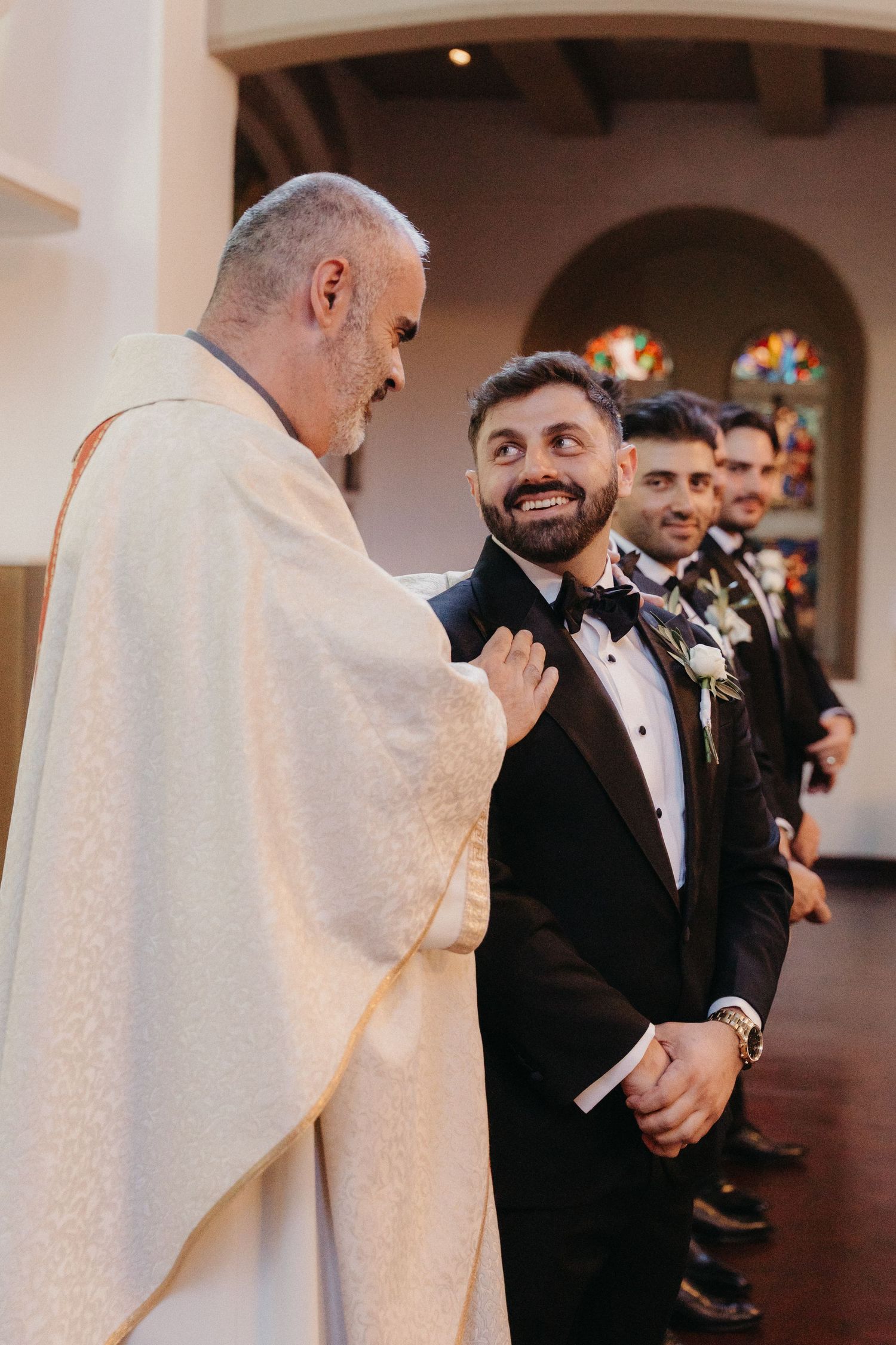 Groom smiling at priest in church wedding ceremony, groomsmen in black tuxedos standing in line behind him.