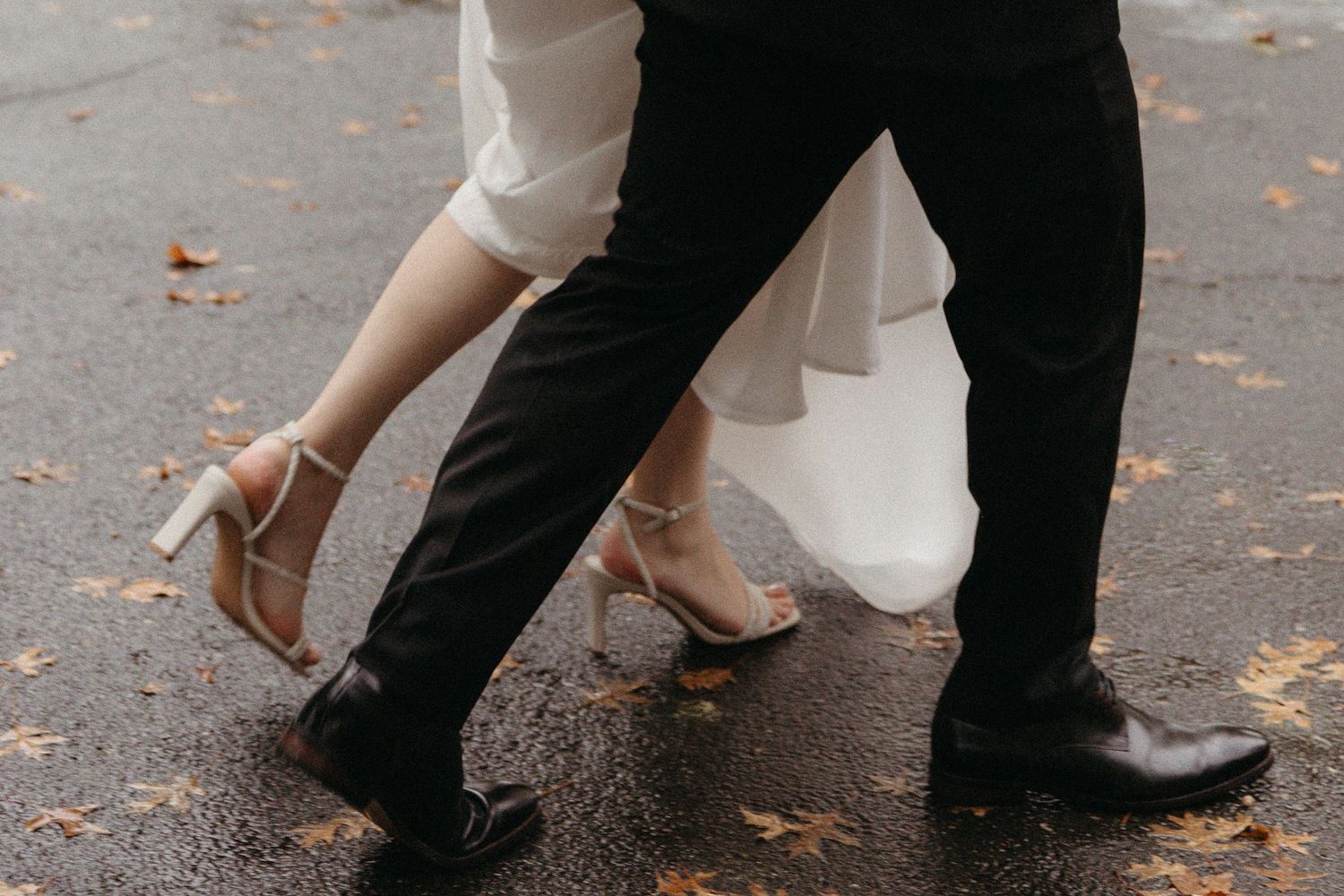 Close-up of bride and groom walking on wet pavement, bride in heeled sandals and white gown, groom in black suit and dress shoes.
