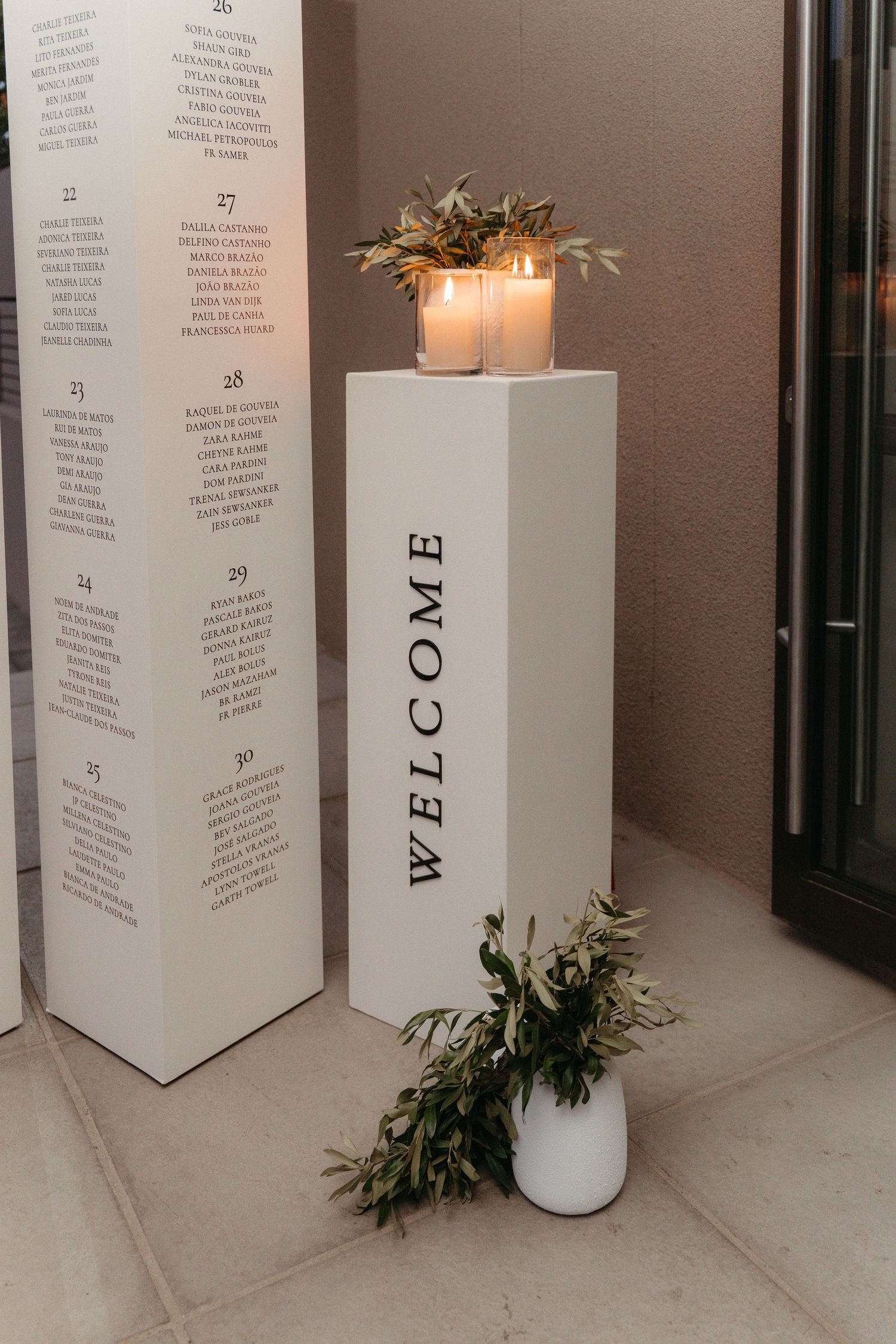 White welcome pedestal with candles and greenery beside a seating chart display at an elegant wedding venue.