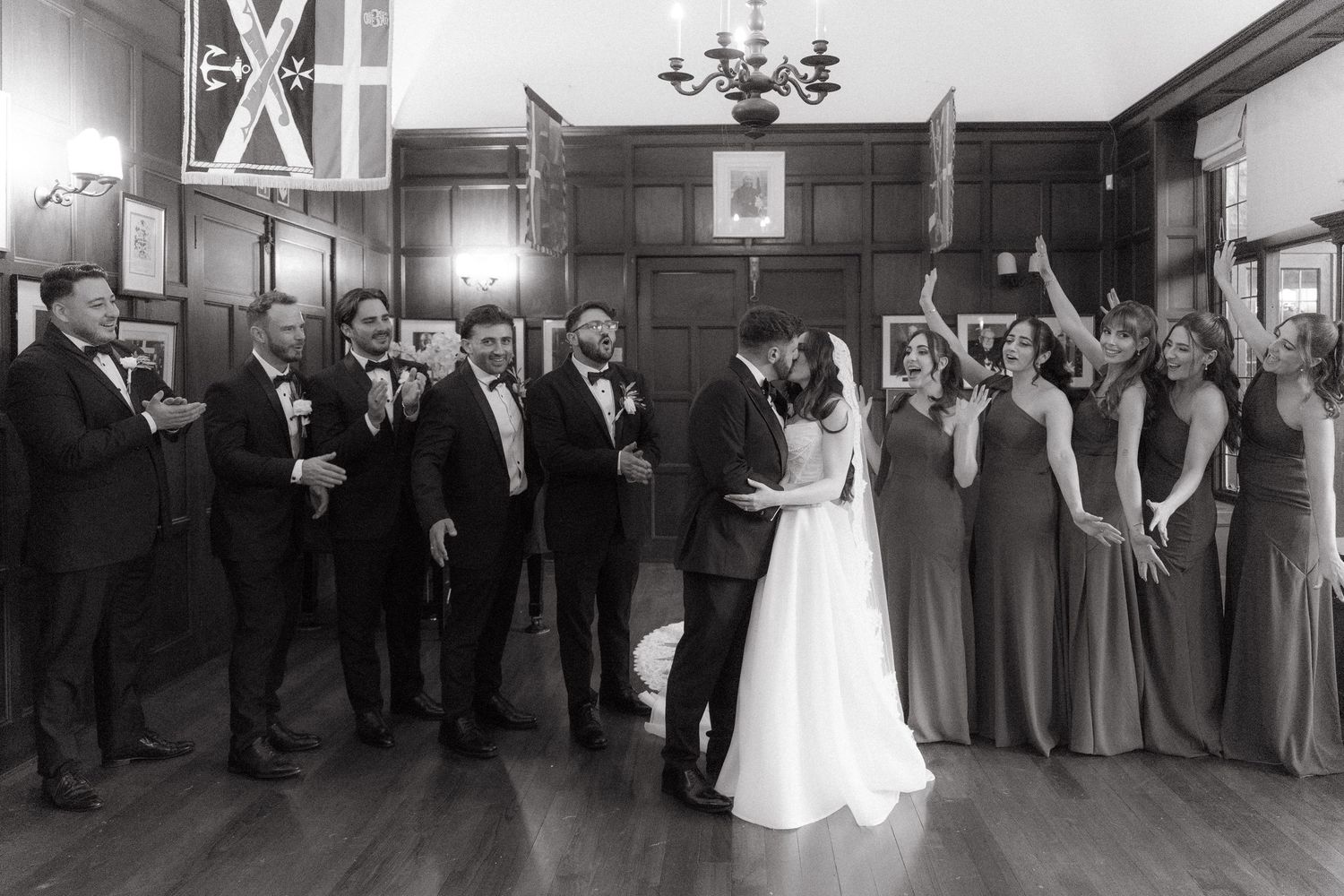 Black and white photo of bride and groom kissing surrounded by cheering wedding party in elegant wood-paneled venue in Johannesburg