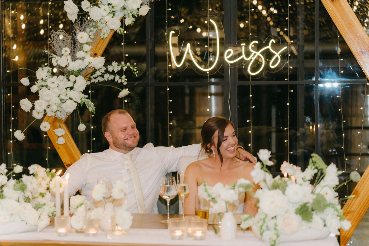 Newlyweds celebrate at reception head table under illuminated 'Weiss' sign and floral decor.