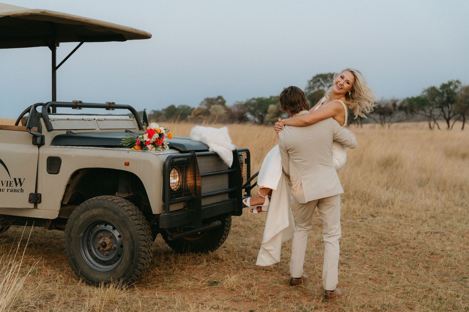Wedding party gathered around a classic vehicle in an open field setting.