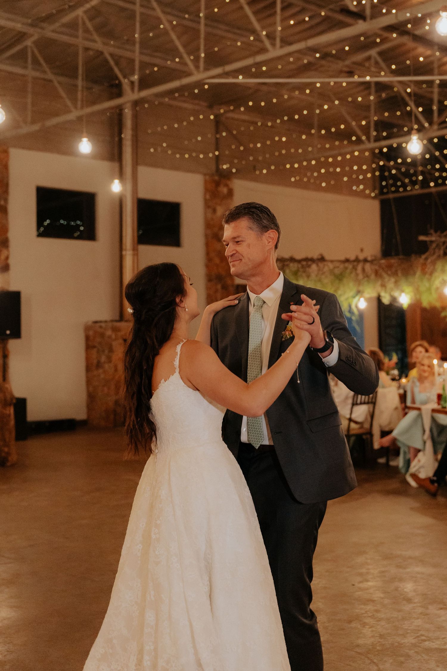 A couple performs their first dance under a canopy of string lights in a spacious reception hall.