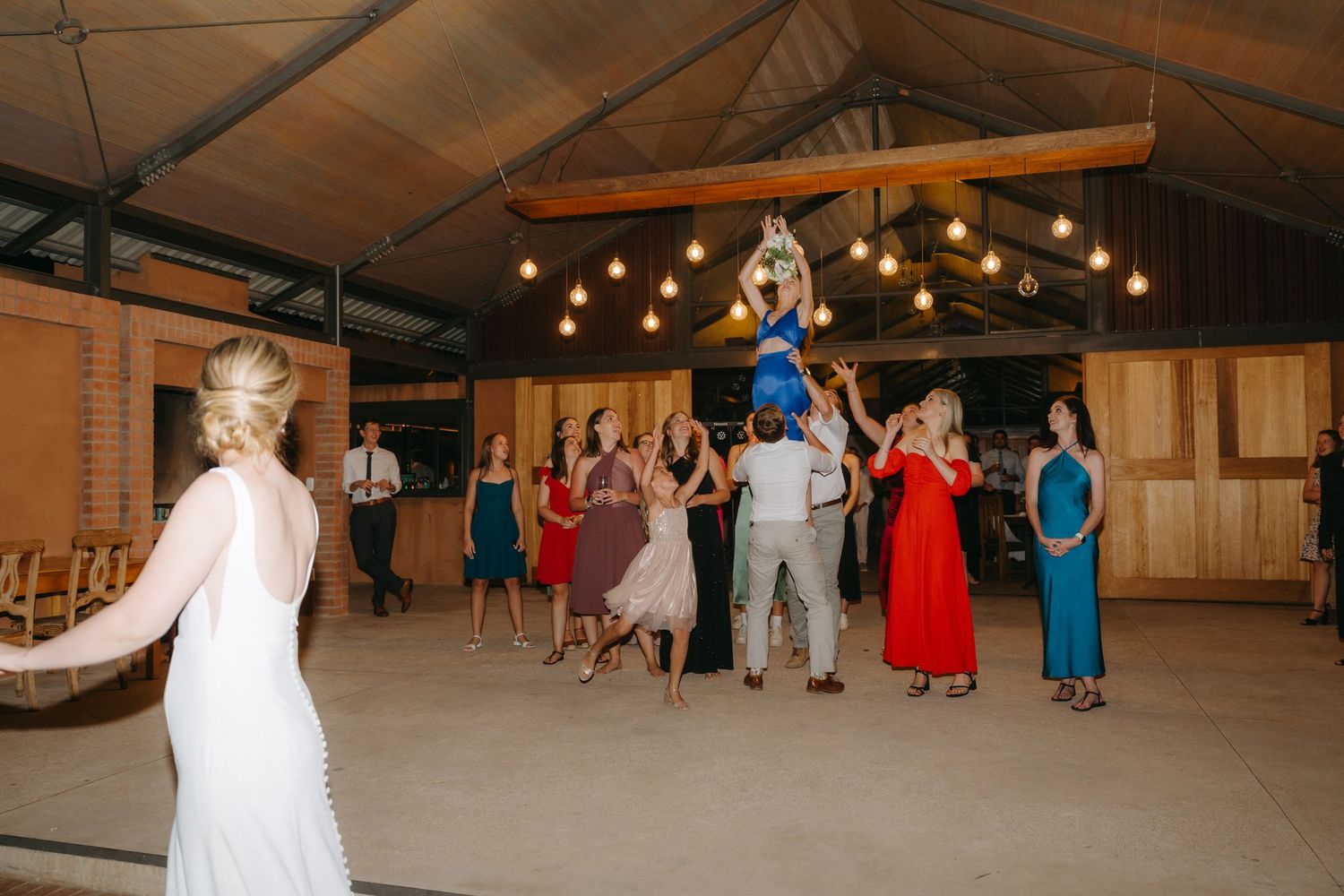 Wedding party guests gather around for the bouquet toss tradition inside a rustic barn reception venue with hanging lights.