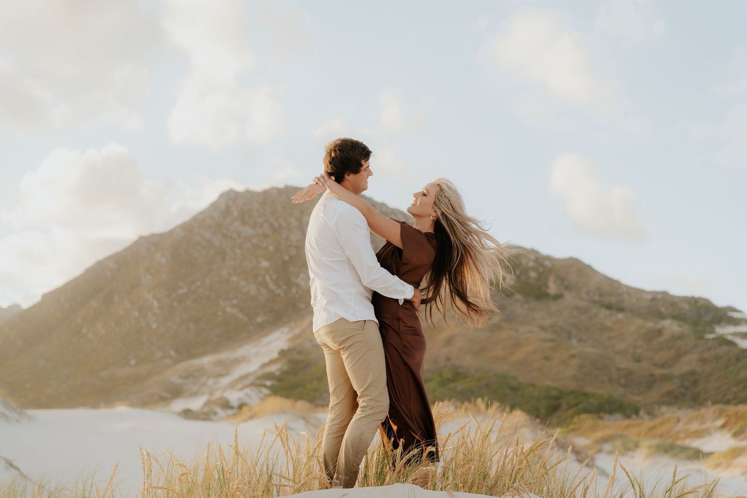 Couple dancing romantically on sandy dunes with mountain backdrop and golden grass in warm sunset light.