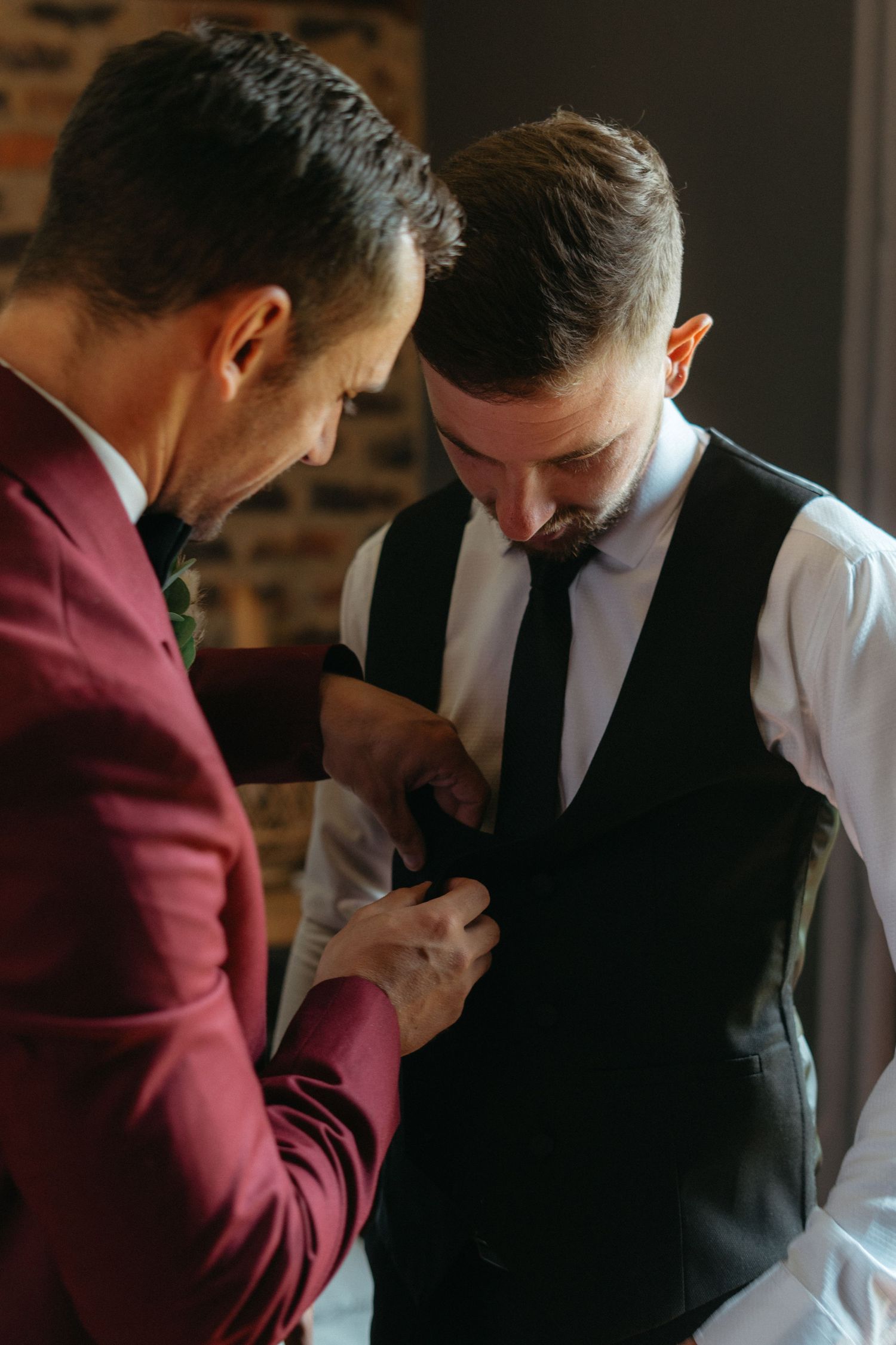 Groom assists in buttoning up another person's vest at a formal wedding preparation.