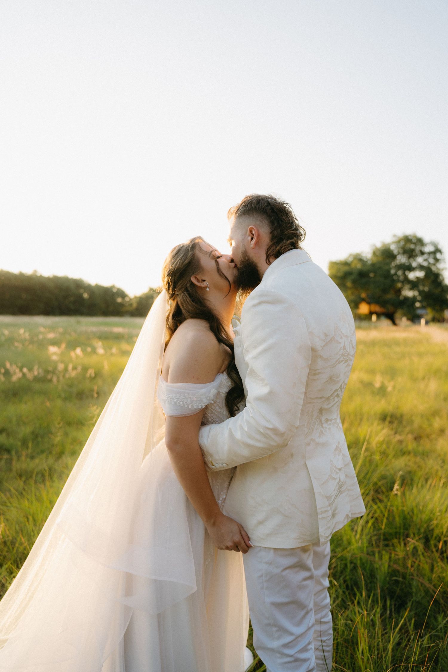 A romantic sunset wedding portrait of a couple in white attire embracing in a golden meadow.
