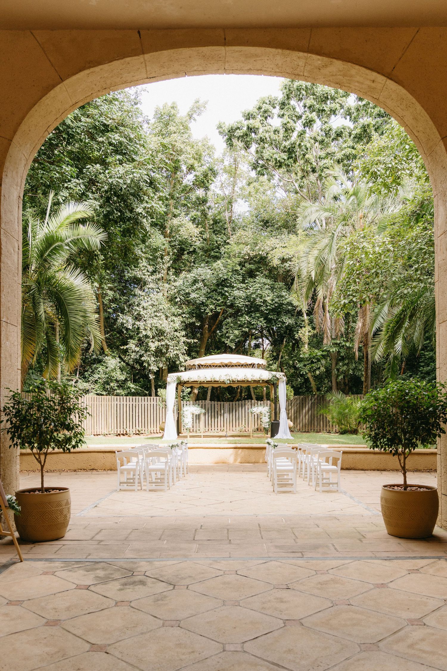 An elegant outdoor wedding ceremony setup with white chairs and arch gazebo viewed through a stone archway with potted plants.