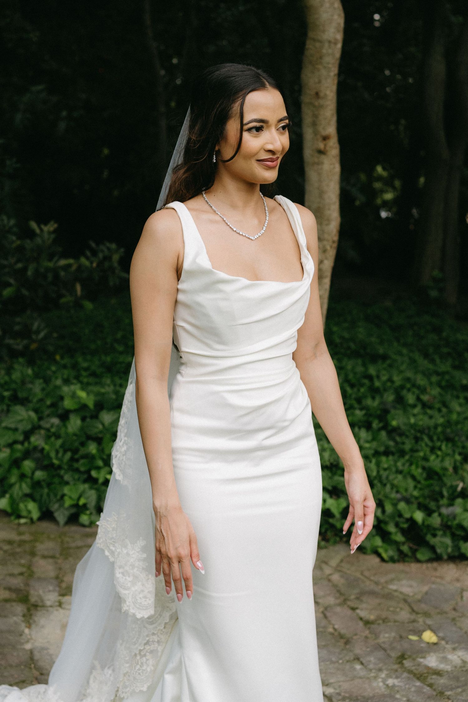 Elegant bride in fitted white satin dress poses in garden setting.