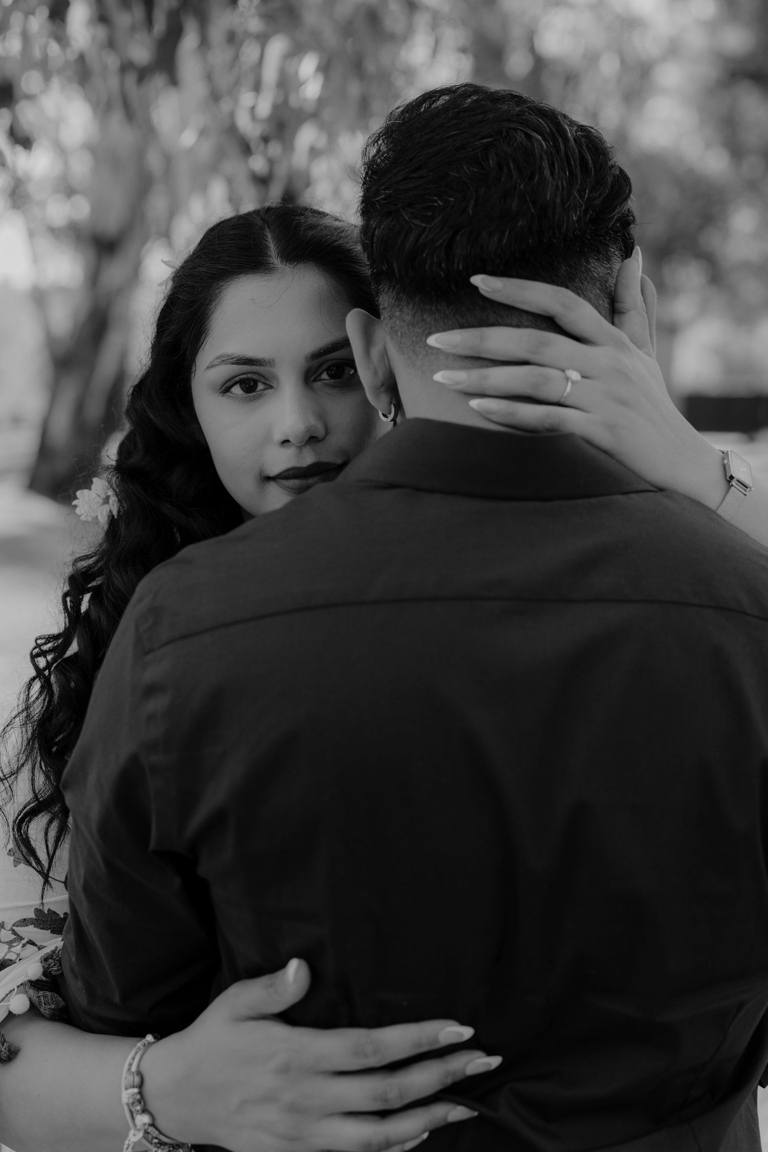 Black and white portrait of couple embracing, showing her hand on his neck with engagement ring visible.