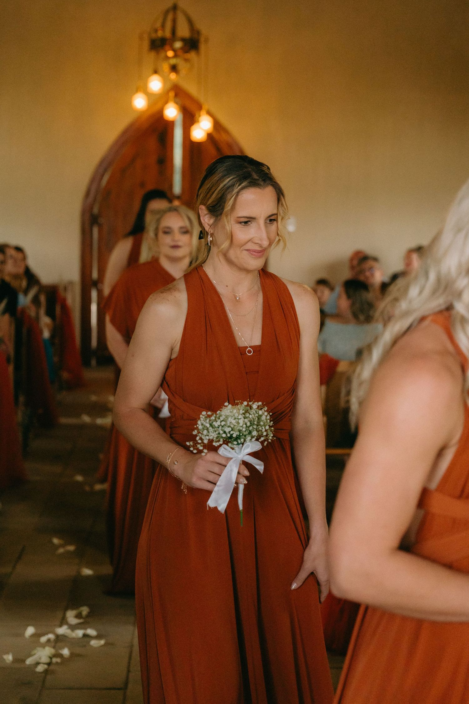 Bridesmaids wearing matching rust-colored gowns hold delicate bouquets during wedding ceremony.