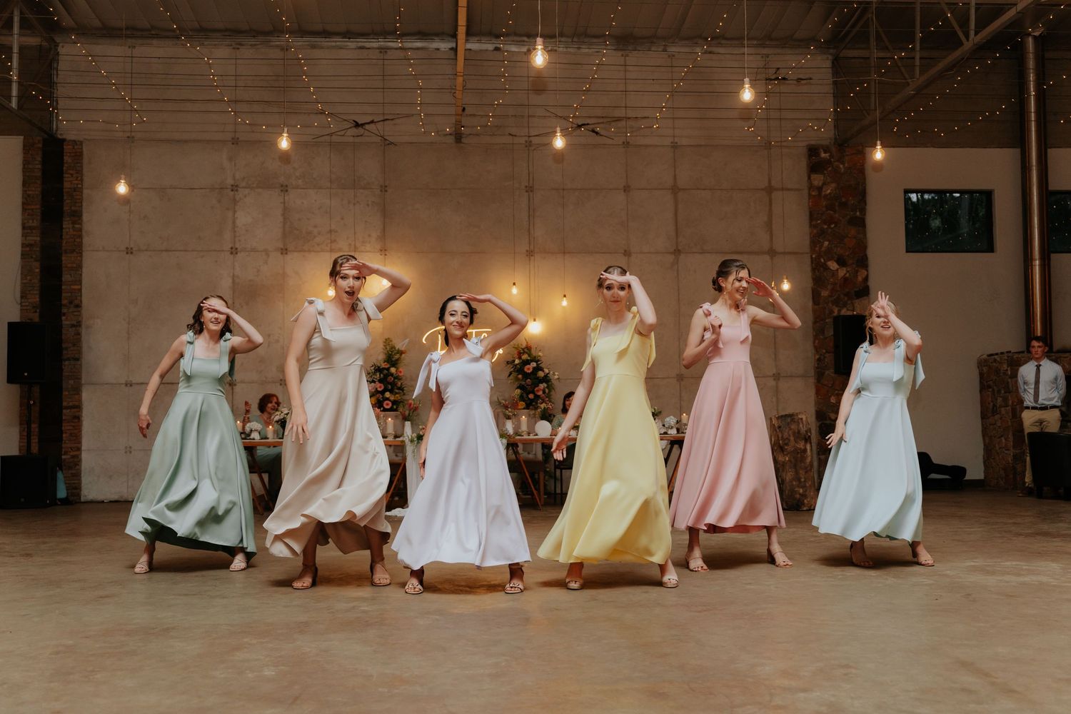 Bridal party in pastel dresses poses together in a rustic warehouse venue with string lights overhead.