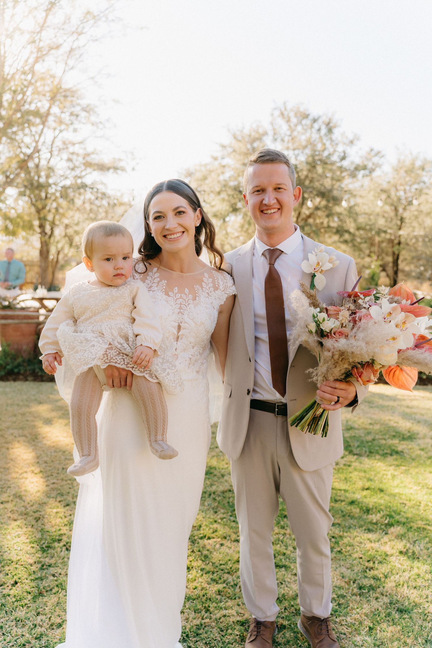 Wedding party members in white and beige attire pose together on a sunny grassy field.