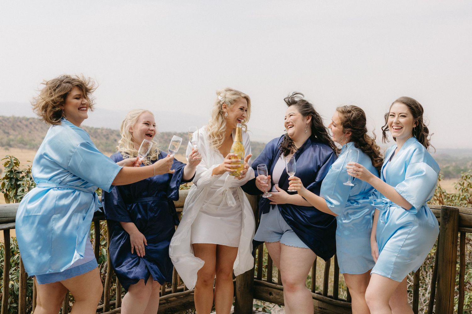 Wedding party laughs together on wooden deck in blue and white attire.