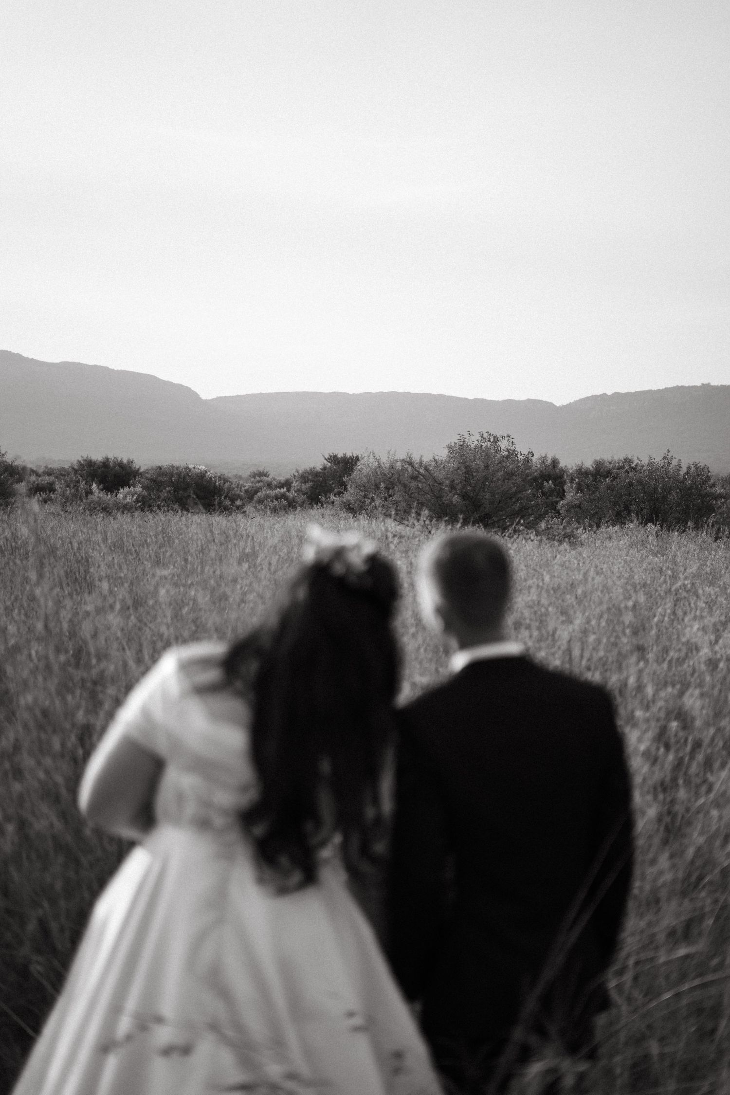A romantic black and white photo of newlyweds standing together in a field with mountains in the distance.