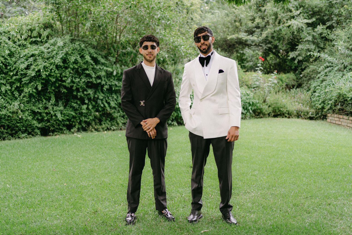 Two men in formal suits pose on manicured lawn at outdoor event.