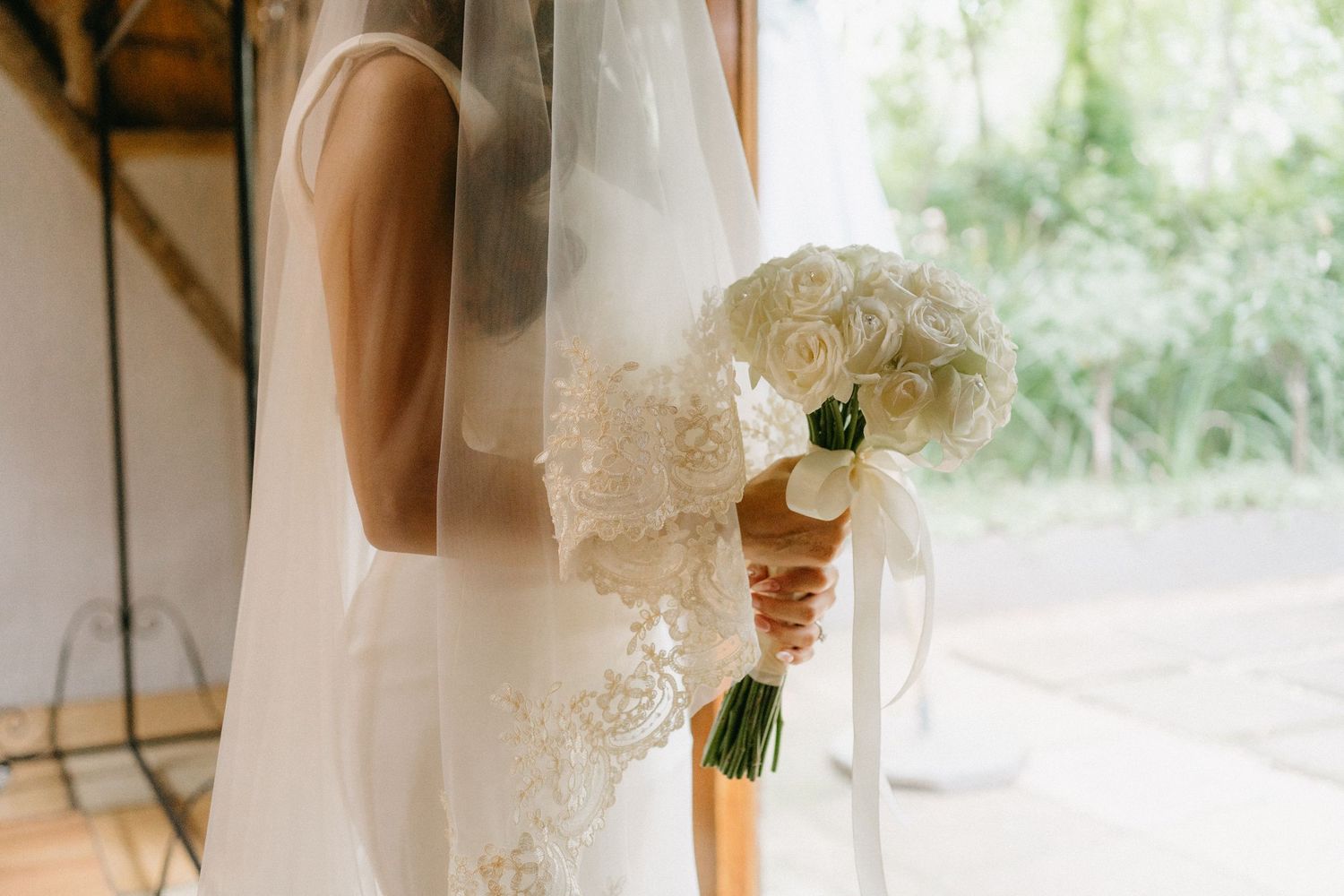 A delicate white bridal bouquet of roses being held against a flowy veil in soft natural light.
