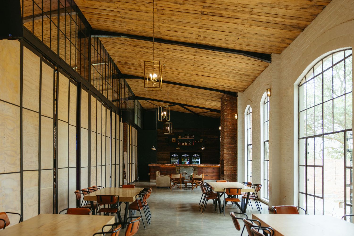 Industrial-style restaurant interior with wooden ceiling, arched windows and minimalist dining setup.