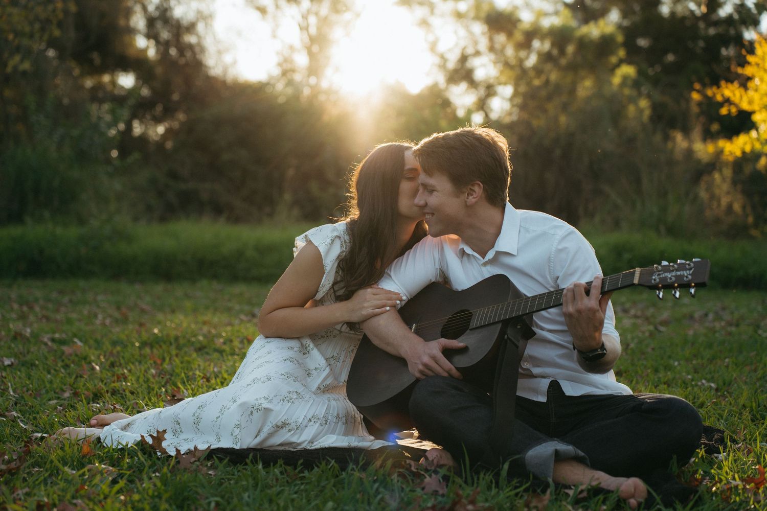 A couple sits together on grass at sunset while one plays acoustic guitar in romantic outdoor setting.