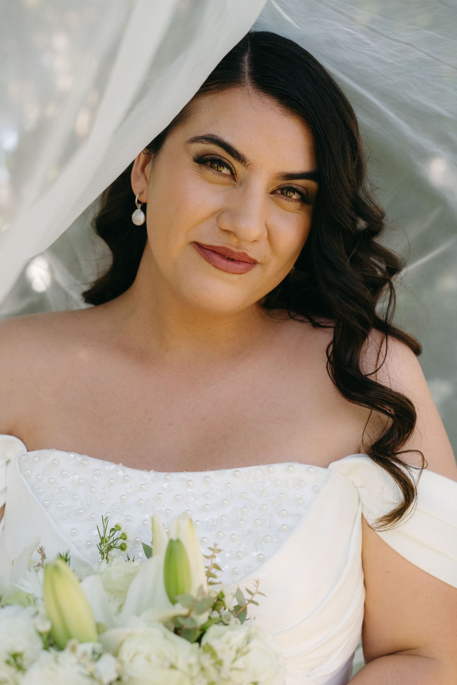 Beautiful bride wearing a white off-shoulder wedding dress and pearl earrings poses with her bridal bouquet of white lilies.