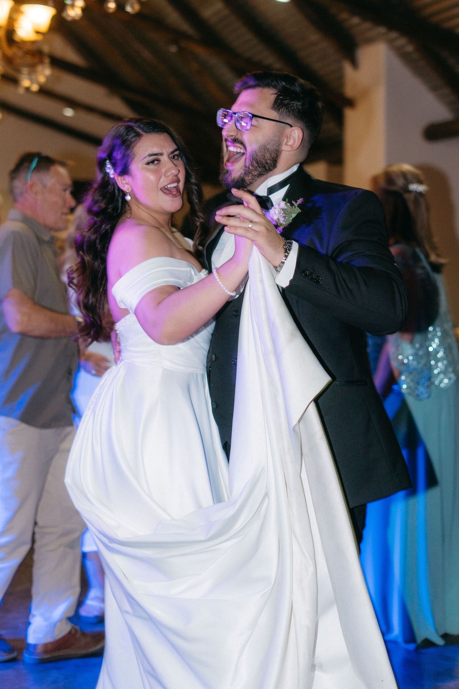 A couple shares their first dance at a wedding reception with blue lighting and exposed wooden beams overhead.