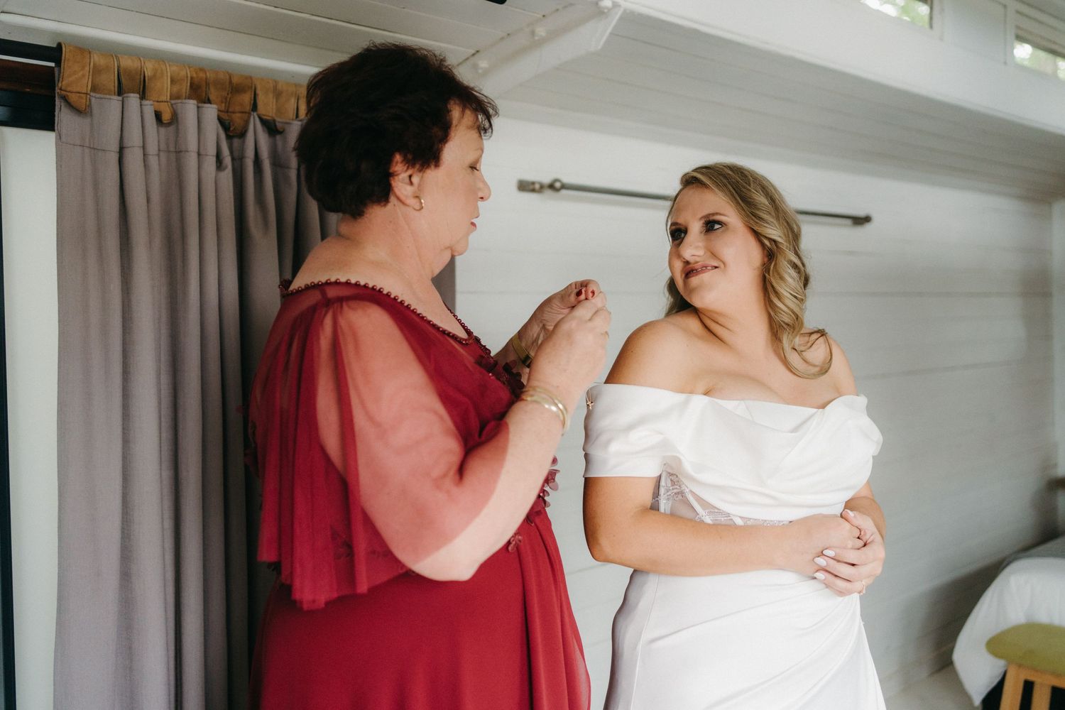 Two people share an intimate moment in a room with curtains while one wears a burgundy dress and another in white.