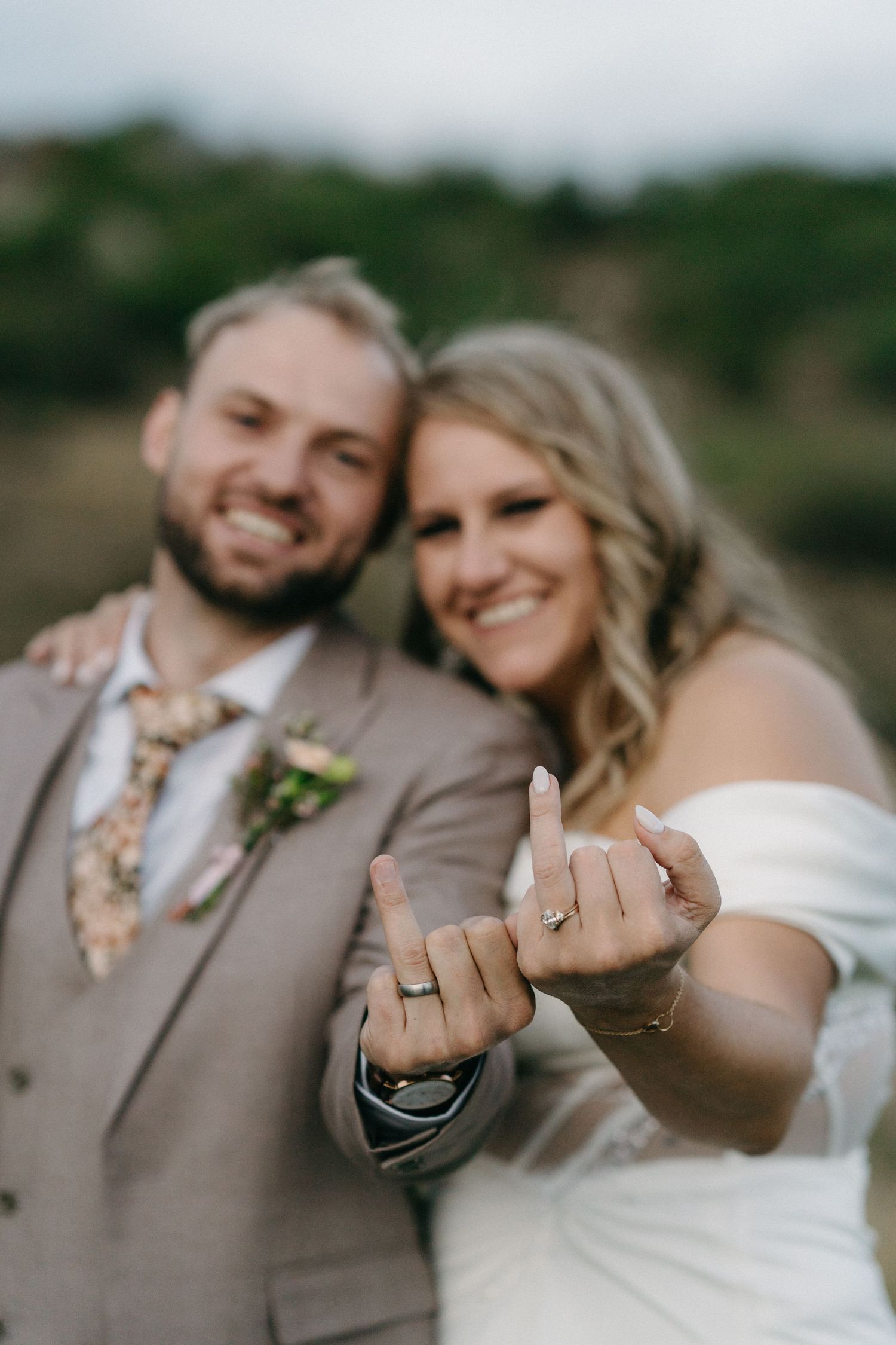 A wedding couple shows off their rings with joyful smiles in an outdoor setting.