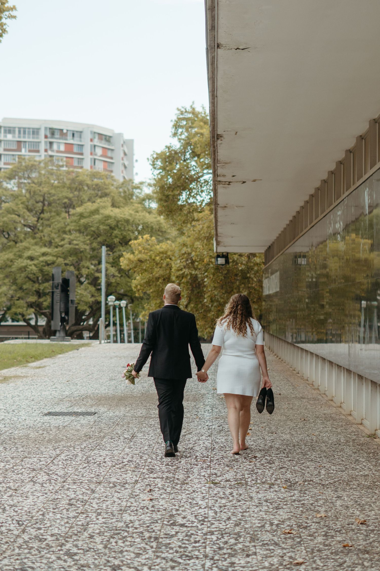 Couple walking hand in hand along path with autumn trees and modern building in background.