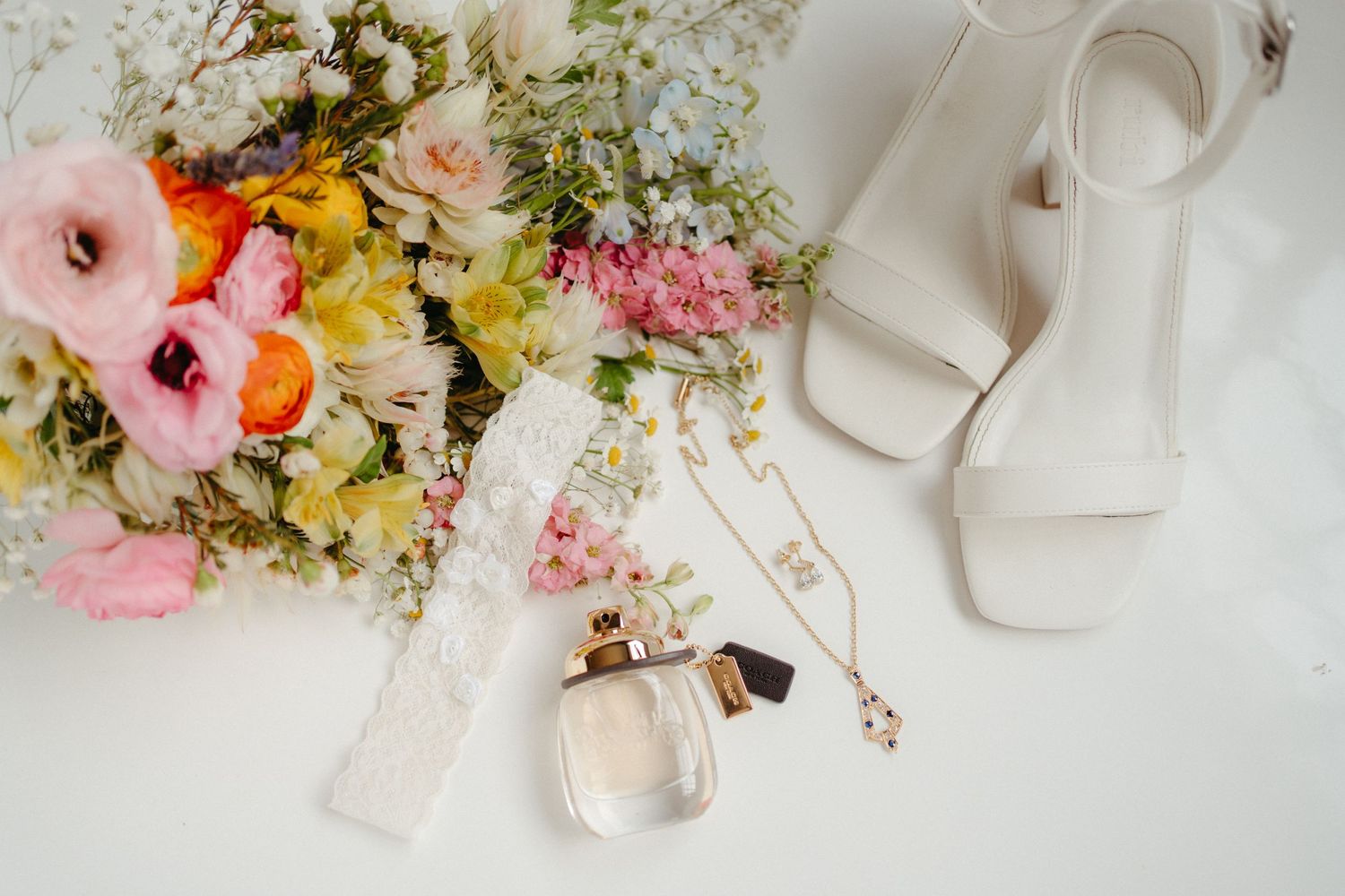 Wedding details flatlay with pink florals, perfume bottle, and white bridal shoes.