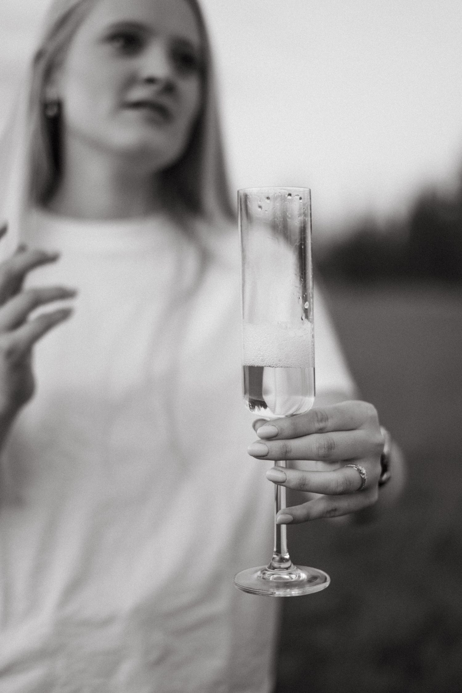 Black and white photo of hand holding champagne flute with engagement ring visible.