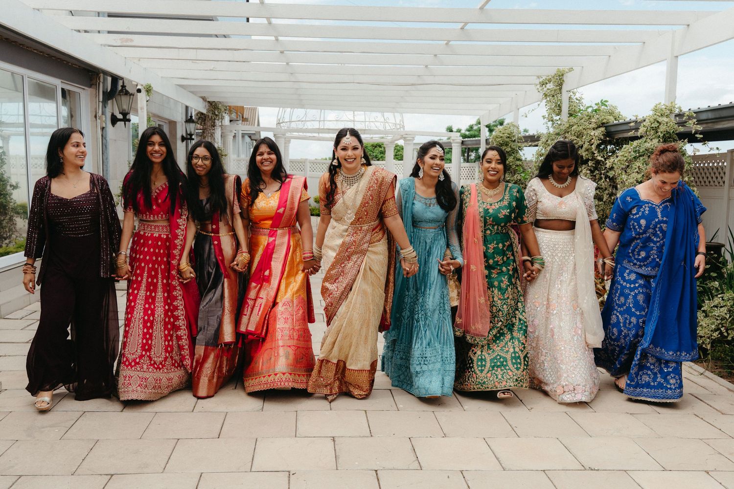 A group of women in colorful traditional Indian saris and formal dresses pose together at an outdoor venue.