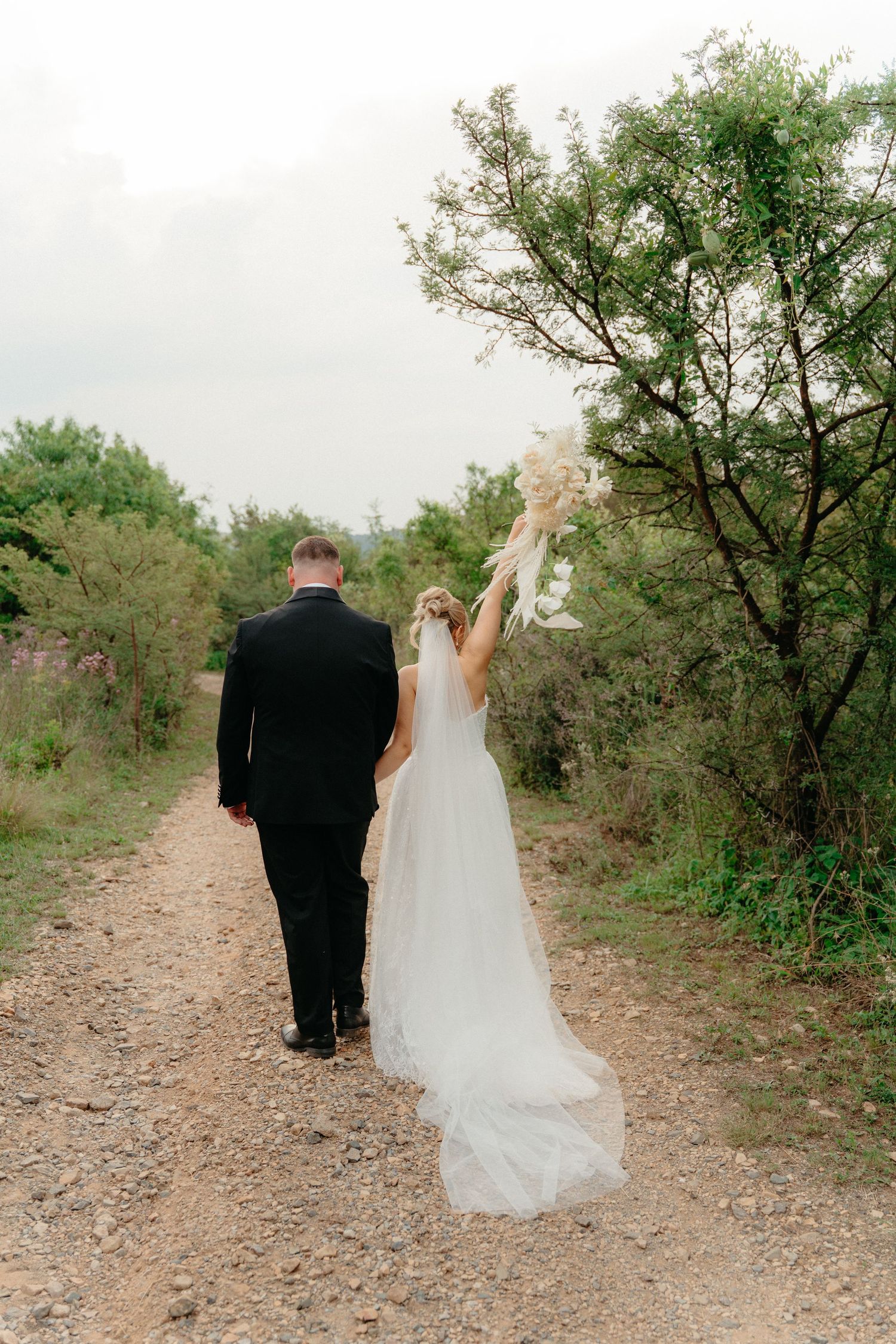 A couple in wedding attire walks along a scenic rocky path surrounded by green trees.