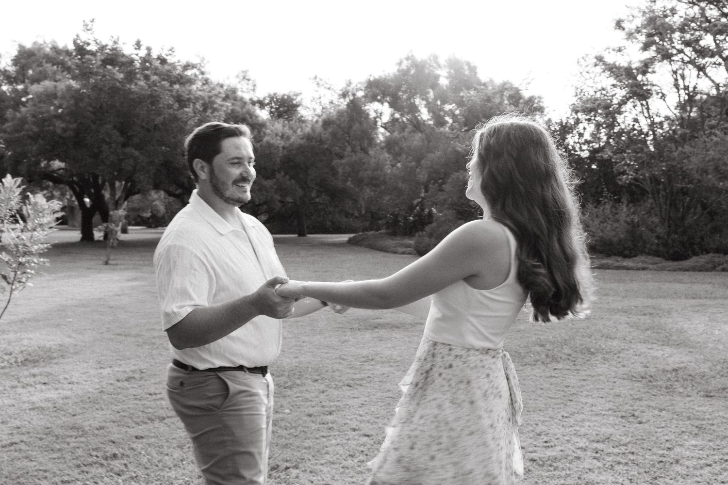 Black and white photo of a couple holding hands and dancing outdoors in a park setting.