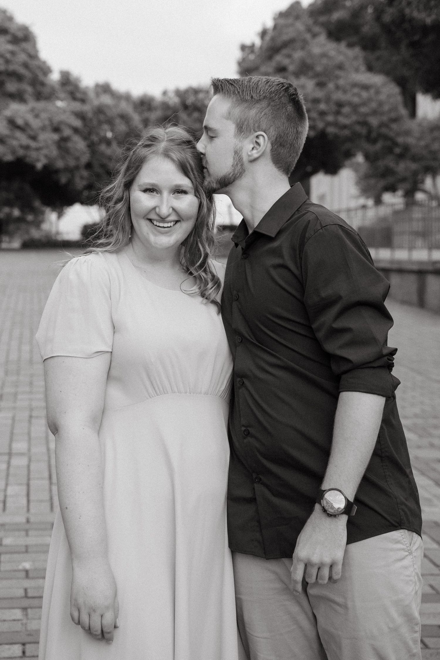 A couple shares a sweet moment together outdoors in black and white, with her in a white dress and him in dark shirt.