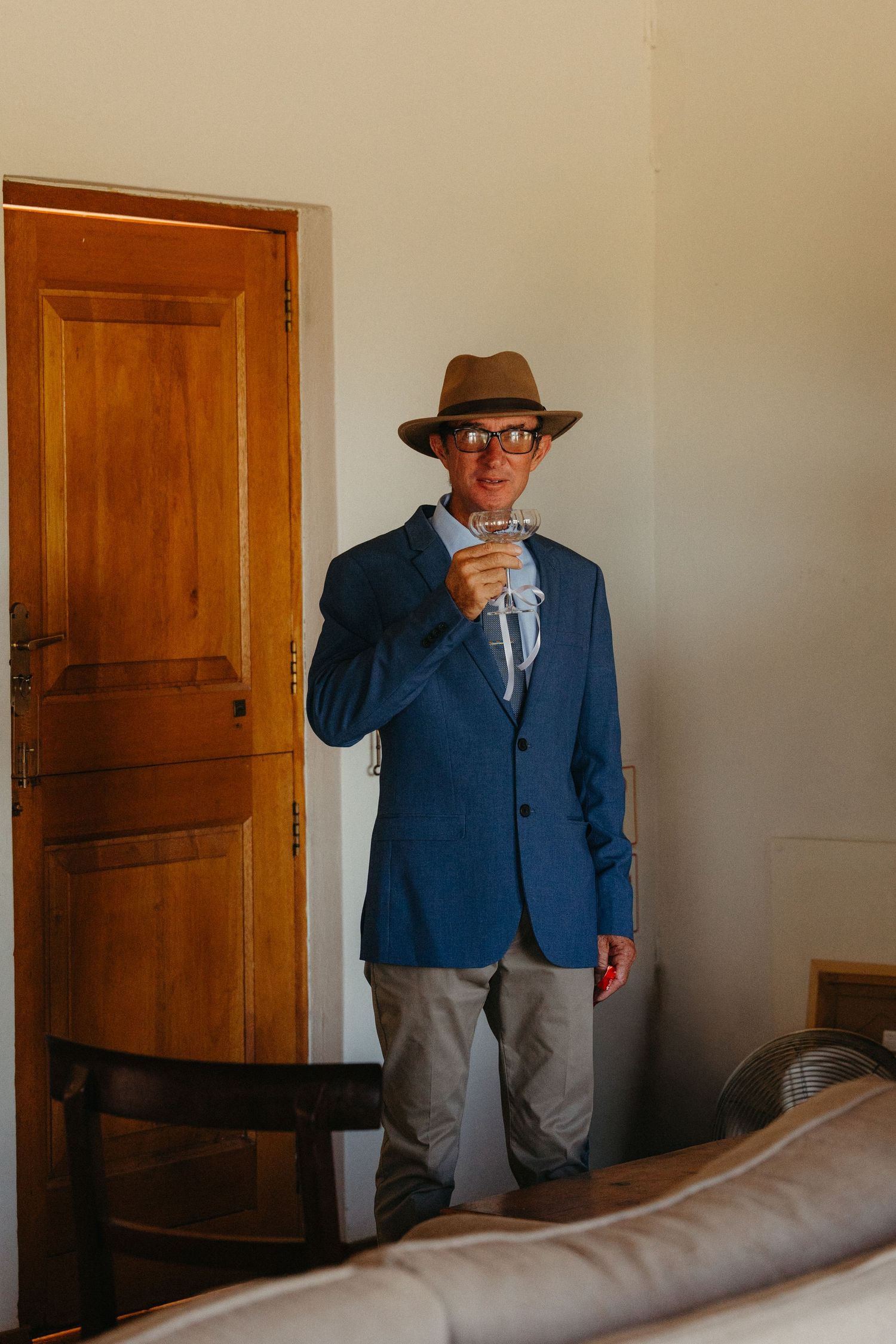 Person in a blue blazer and brown hat stands in a corner by wooden doors in a minimalist interior space.